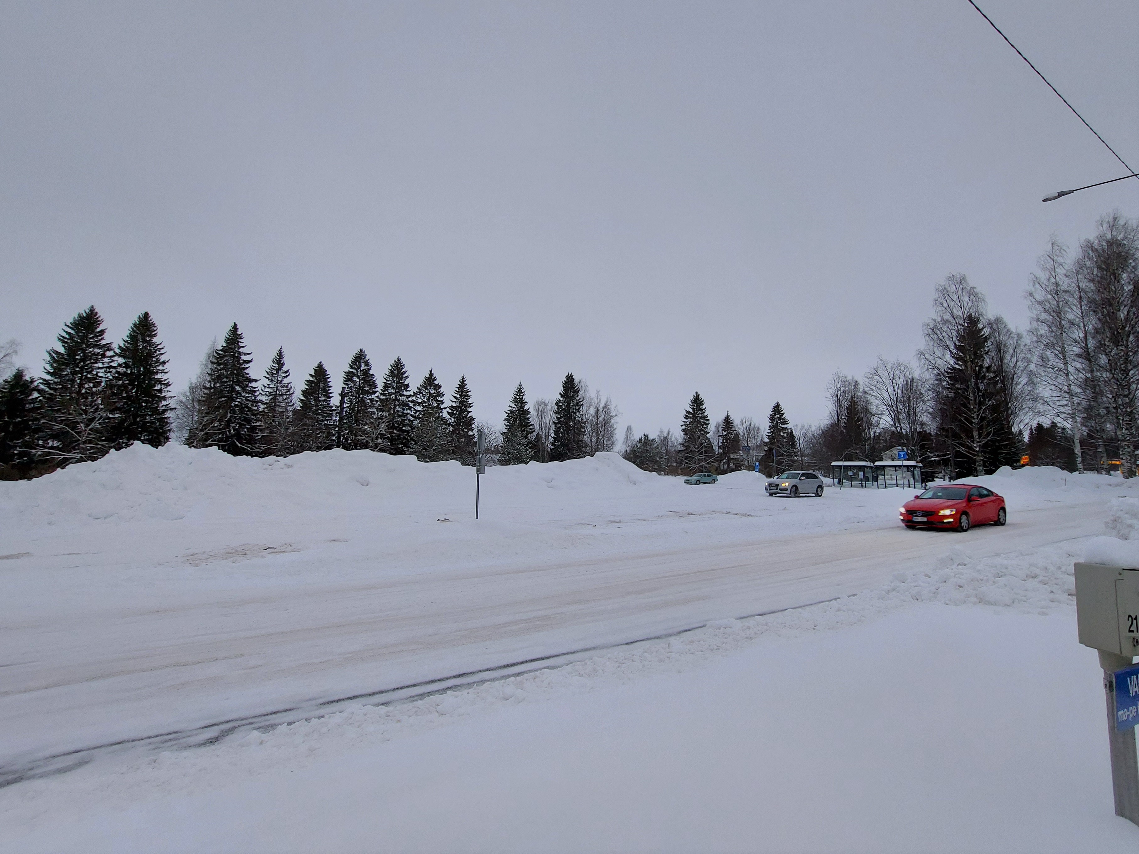 Talvinen maisema, isoja lumikasoja. Kaksi autoa, toinen vaalea toinen punainen.