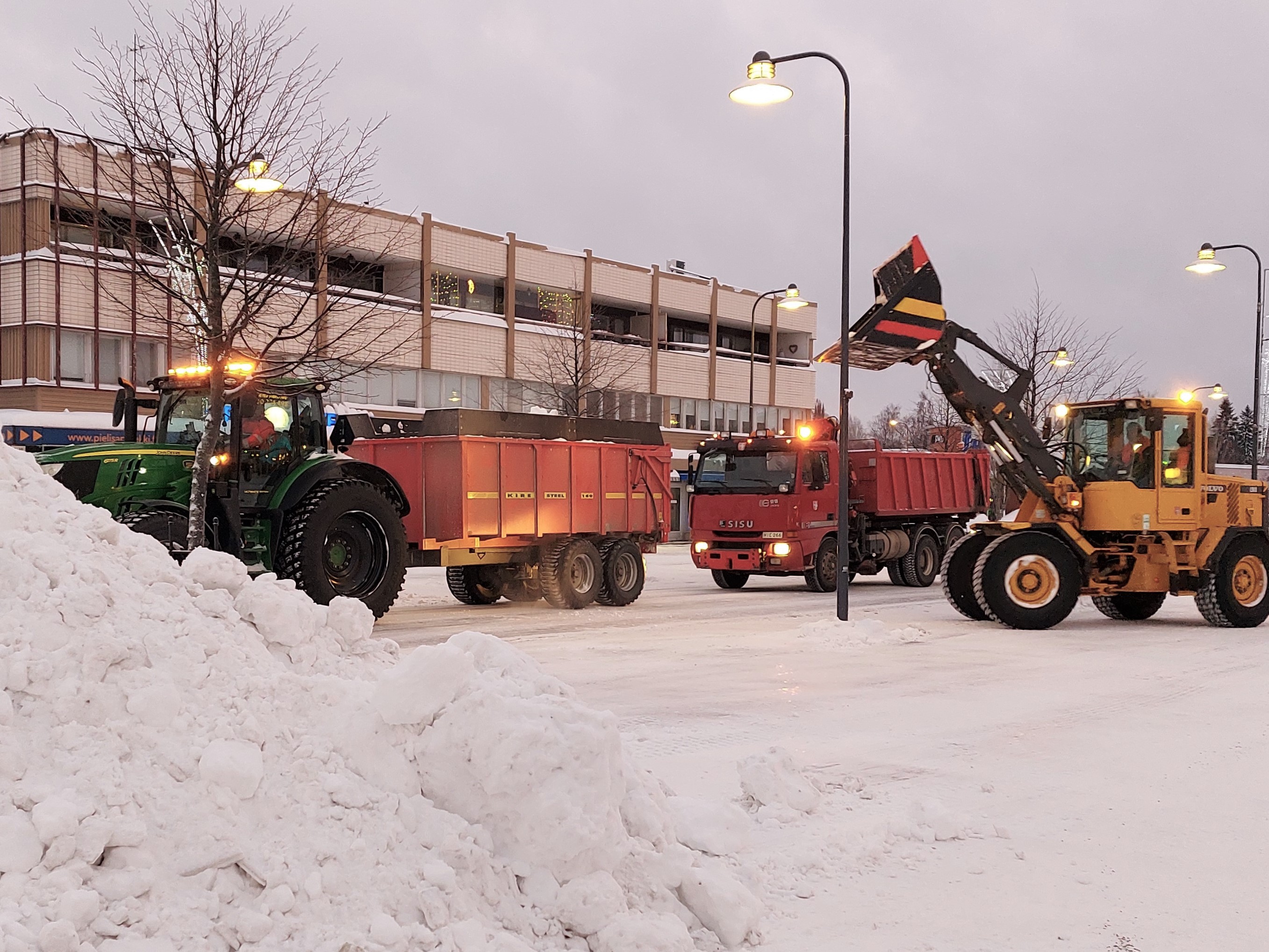 Lumikasan takana traktori ja kuorma-auto. Kauhakuormaaja lastaa lunta auton kyytiin.