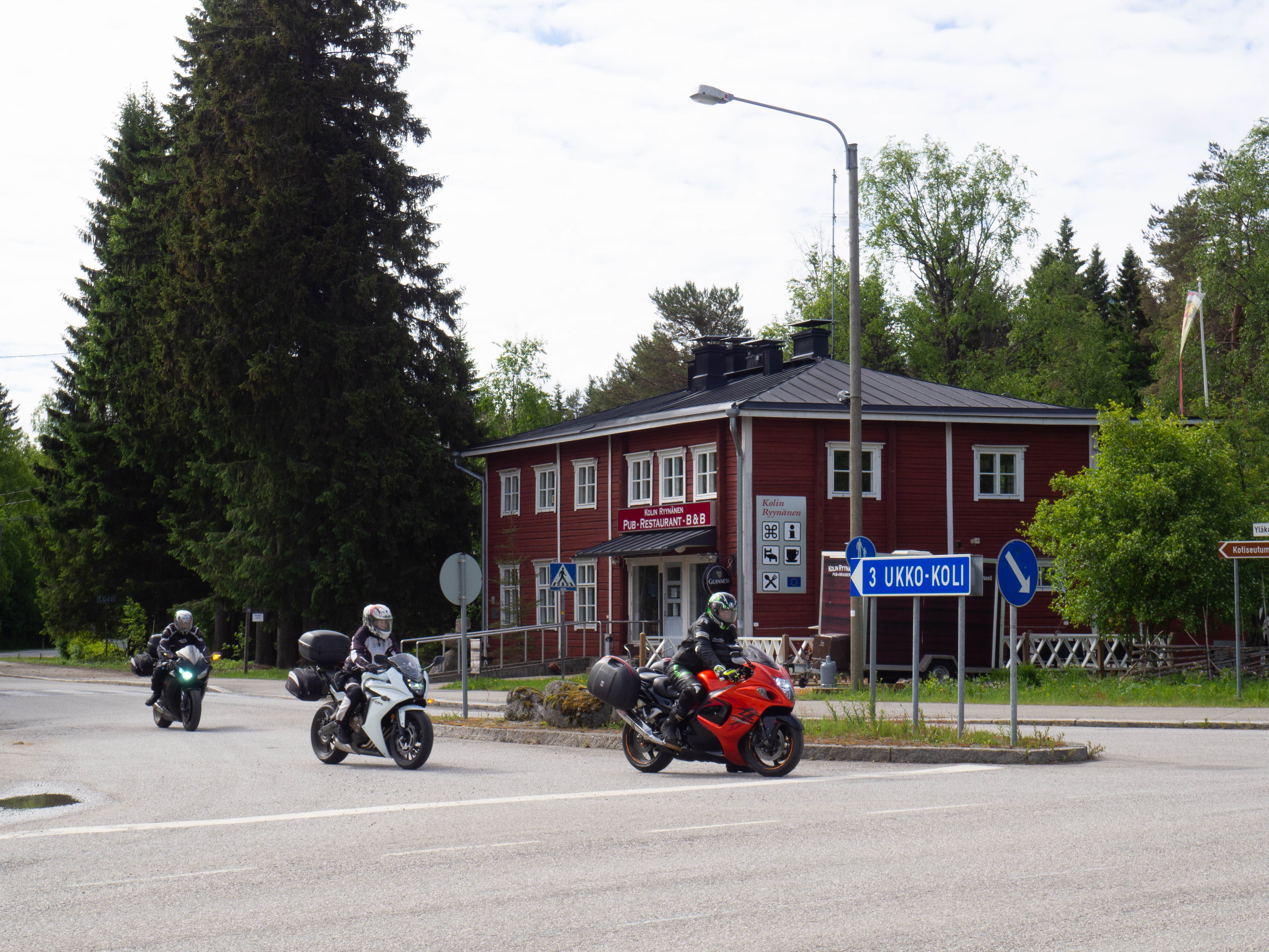 Kolme moottoripyöräilijää risteyksessä. Tienviitassa teksti Ylä-Koli 3. Taustalla iso punainen hirsirakennus.