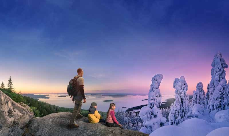 Kolin maisema, joka vaihtuu kuvassa kesästä talveen. Kolme ihmistä kalliolla katsomassa maisemaa. 