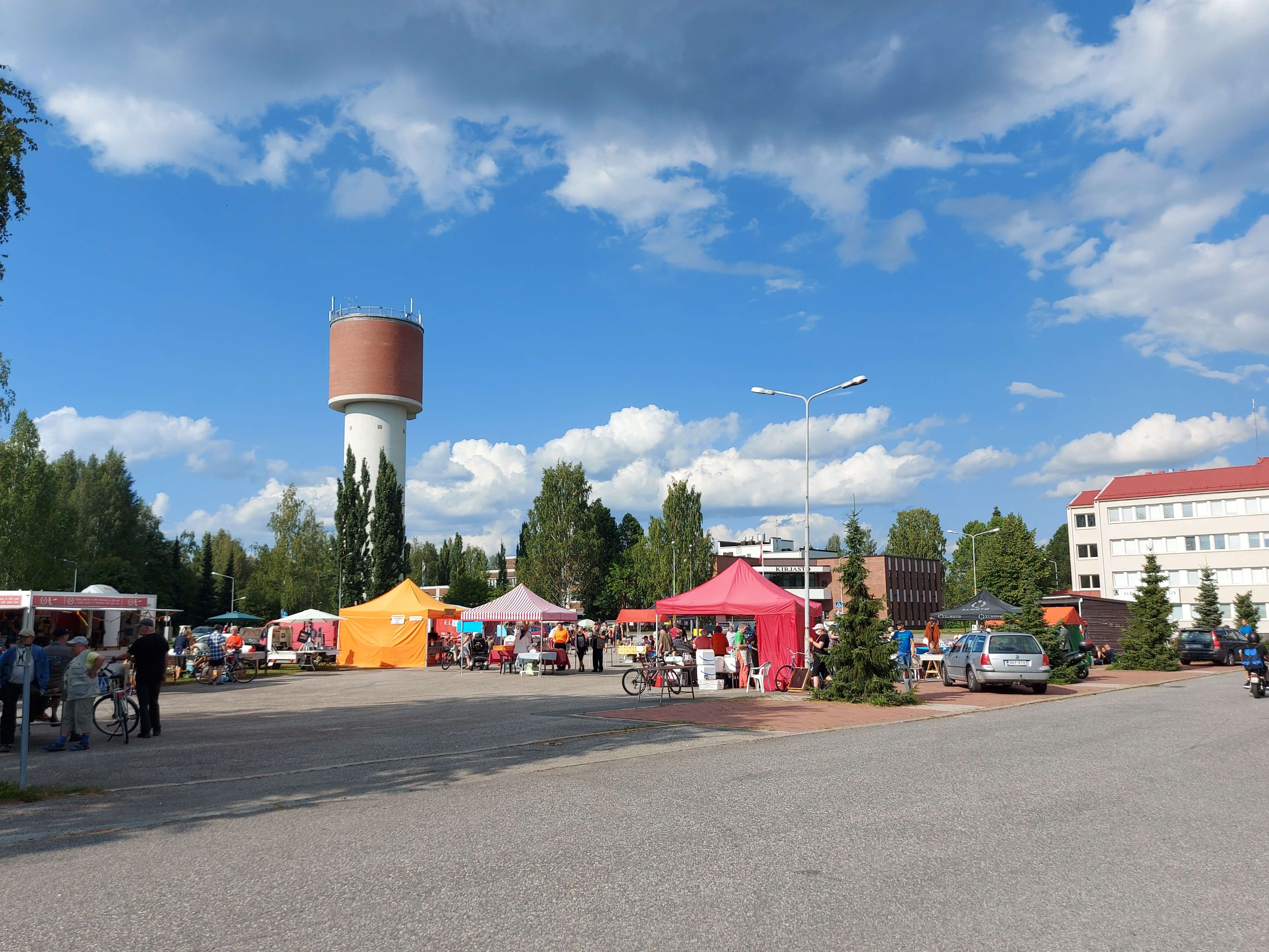 Lieksan torilla myyntikojuja ja niiden väleissä ihmisiä. Taustalla korkea vesitorni ja kaupungin kirjasto. 