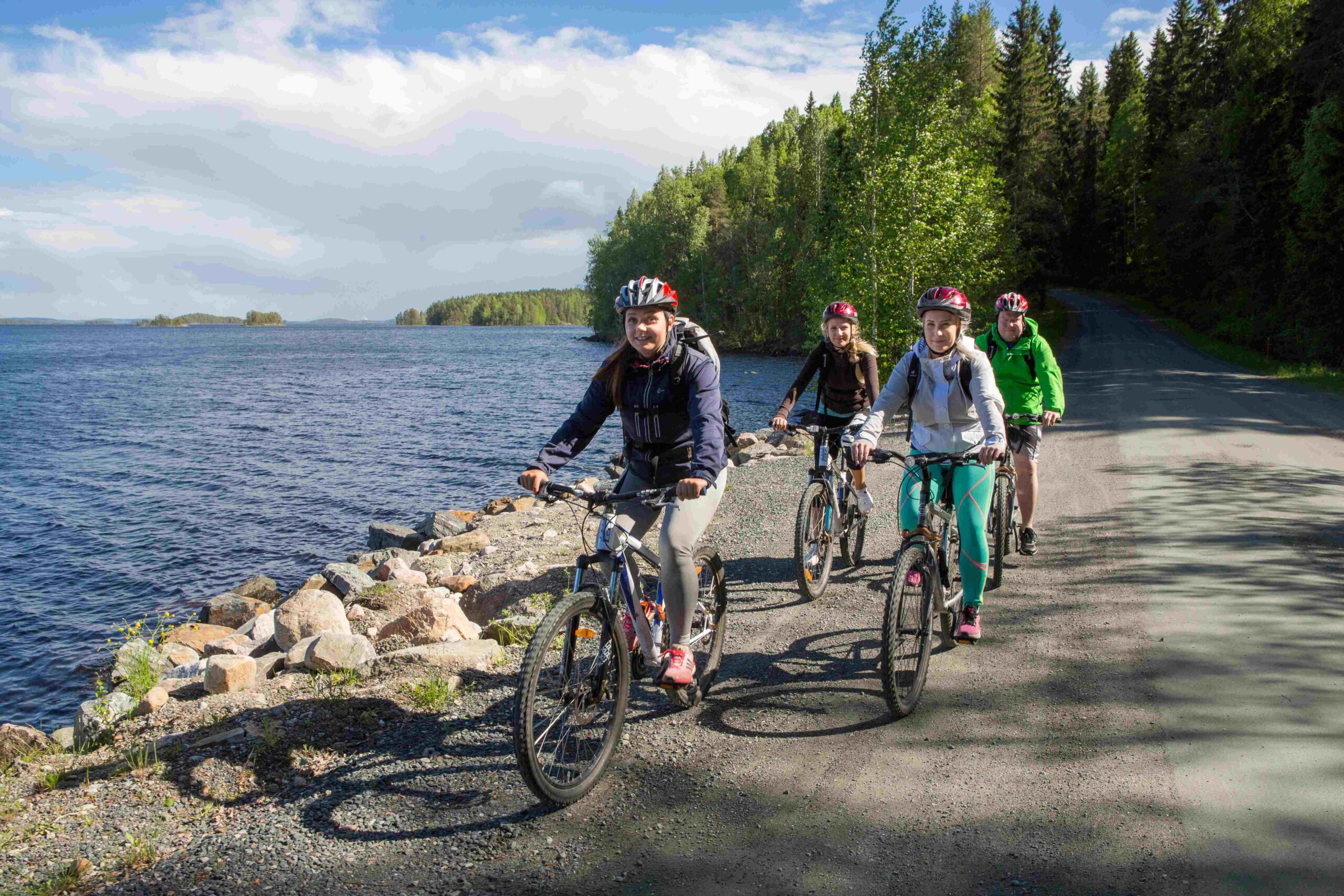 Neljä pyöräilijää ajamassa polkupyörillä tien reunassa. Tie kulkee rantaa pitkin, tien vieressä on järvi.