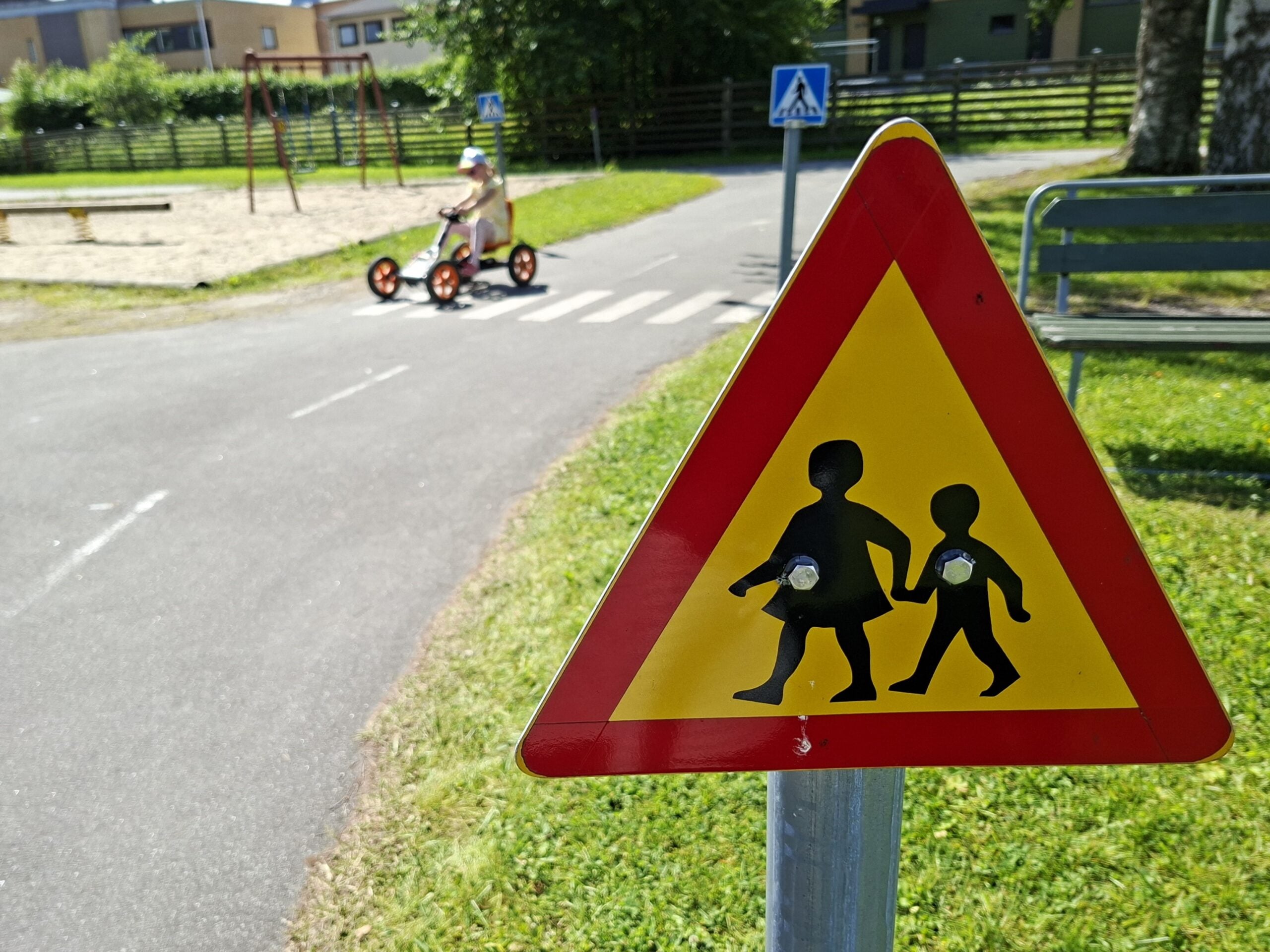 Liikennemerkki, joka varoittaa lapsista liikenteessä. Kauempana lapsi ajamassa polkuautolla.